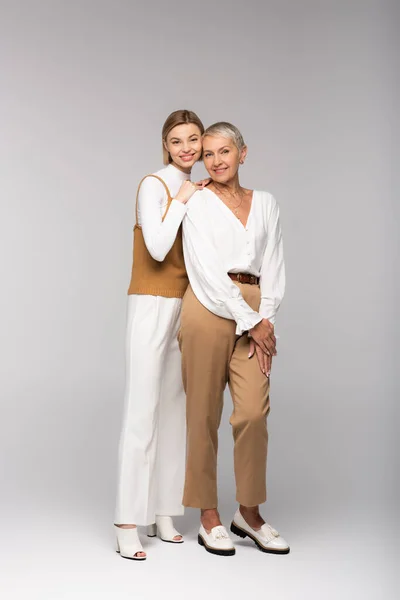 Longitud completa de complacida hija joven abrazando alegre de mediana edad madre en gris — Stock Photo