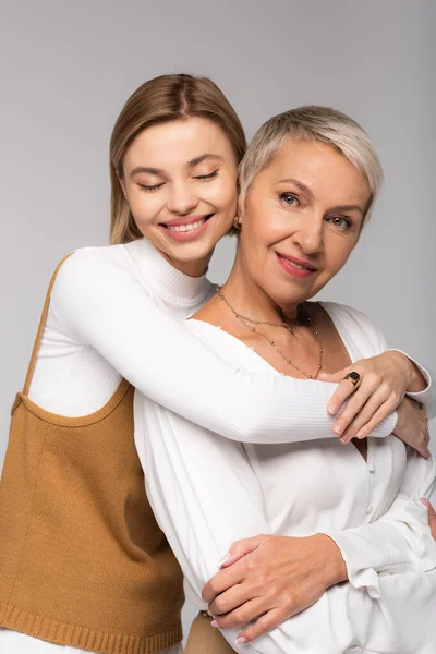 Complacida hija joven abrazando a la madre de mediana edad aislada en gris - foto de stock