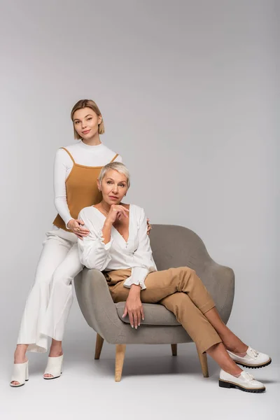 Full length of young woman sitting near mother in armchair on grey — Stock Photo