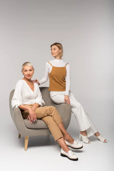 Pleine longueur de jeune femme assise près de la mère joyeuse dans le fauteuil sur gris — Photo de stock