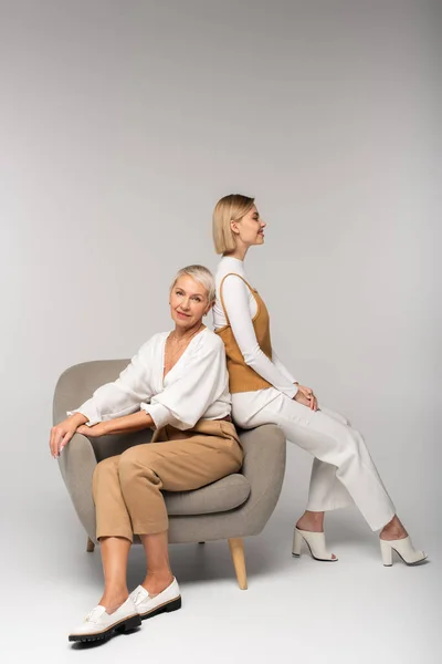 Full length of happy young woman sitting near pleased mother in armchair on grey — Stock Photo