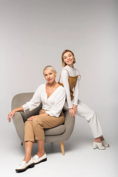 Повна довжина позитивної молодої жінки, що сидить біля матері в кріслі на сірому — стокове фото
