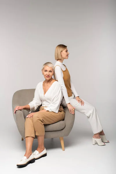 Longitud completa de la joven sentada cerca de la madre positiva en el sillón en gris — Stock Photo