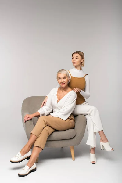 Full length of young woman sitting near smiling middle aged mother in armchair on grey — Stock Photo