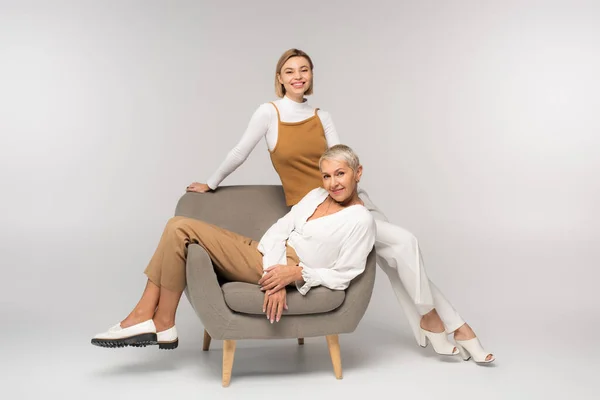 Full length of cheerful young woman near smiling mother sitting in armchair on grey — Stock Photo