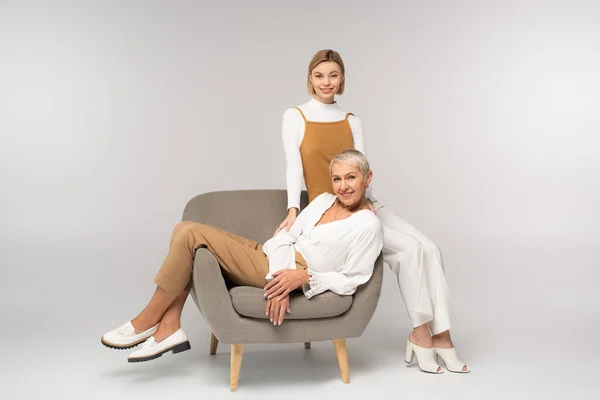 Full length of cheerful young woman near positive mother sitting in armchair on grey — Stock Photo