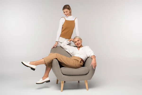 Full length of cheerful young woman standing near smiling mother sitting in armchair on grey — Stock Photo