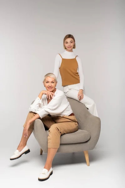 Full length of joyful young woman near smiling mother sitting in armchair on grey — Stock Photo