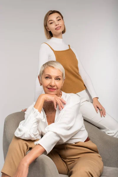 Joyful young woman near smiling mother sitting in armchair isolated on grey — Stock Photo