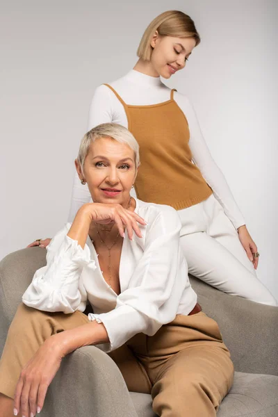 Feliz joven mujer cerca sonriente madre sentado en sillón aislado en gris - foto de stock