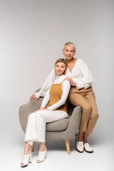 Full length of positive young daughter sitting in armchair near smiling mother on grey — Stock Photo