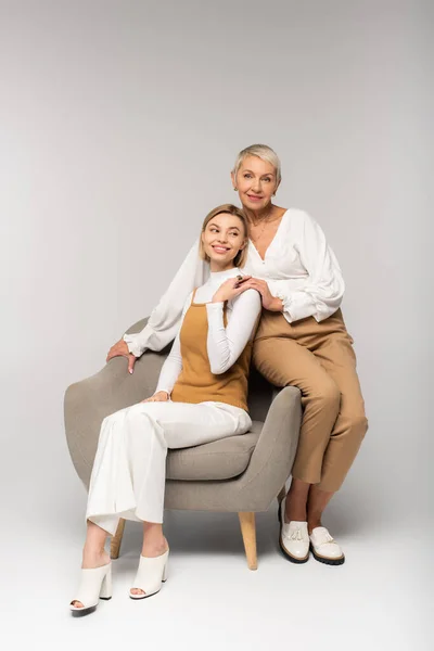 Comprimento total da jovem filha feliz sentada em poltrona perto de mãe sorridente no cinza — Stock Photo