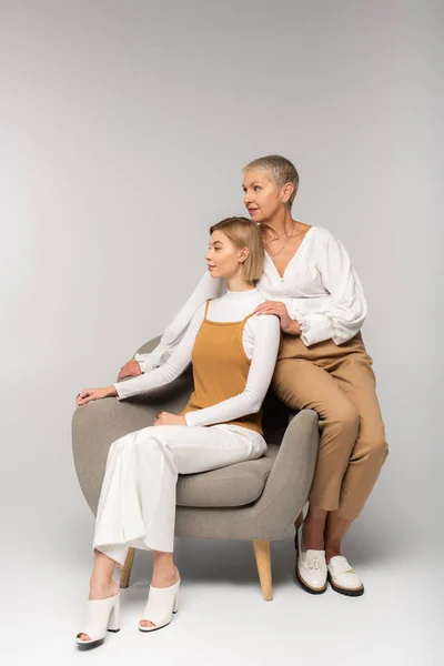Volle Länge der kleinen Tochter sitzt im Sessel neben Mutter mittleren Alters auf grau — Stockfoto