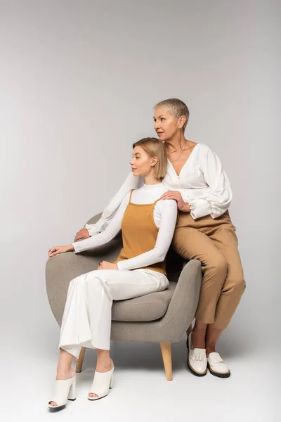 Full length of young daughter sitting in armchair near middle aged mother on grey — Stock Photo