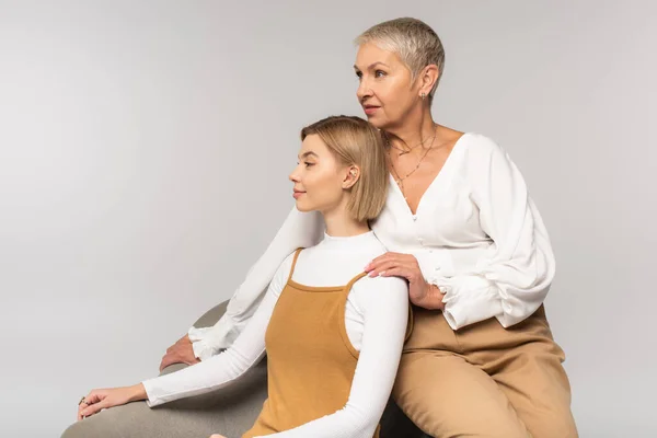 Hija joven sentada en sillón con madre de mediana edad aislada en gris - foto de stock