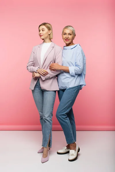 Piena lunghezza di elegante giovane donna e felice madre matura in jeans e camicetta in piedi su rosa — Foto stock