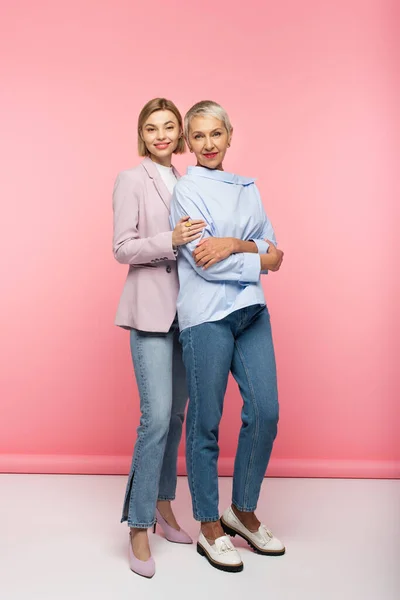 Comprimento total de alegre jovem mulher abraçando mãe madura em jeans e blusa, enquanto em pé sobre rosa — Fotografia de Stock