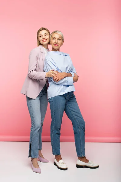 Longitud completa de la joven feliz abrazando a la madre madura en jeans y blusa mientras está de pie en rosa - foto de stock