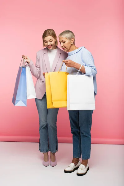 Longitud completa de la mujer feliz y madre madura en jeans mirando bolsas de compras en rosa - foto de stock