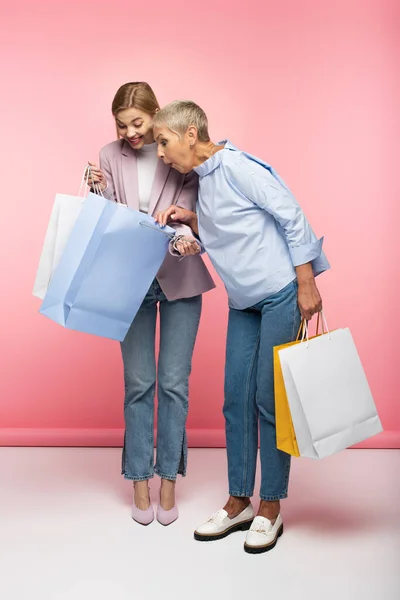 Pleine longueur de femme gaie et surprise mère mature en jeans regardant des sacs à provisions sur rose — Photo de stock