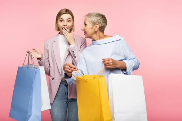 Étonné jeune femme couvrant la bouche près de la mère mature tenant des sacs à provisions isolés sur rose — Photo de stock