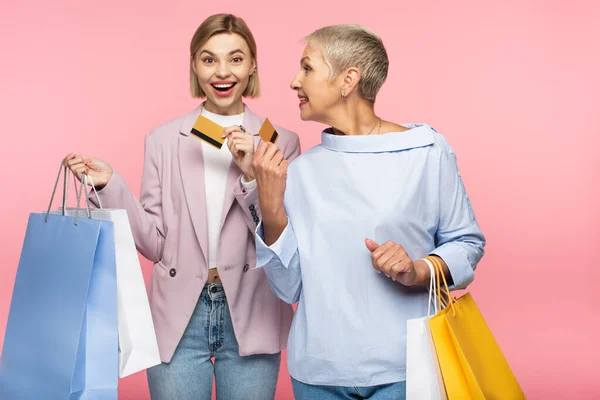 Feliz joven y madura madre sosteniendo bolsas de compras y tarjetas de crédito aisladas en rosa - foto de stock