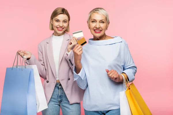 Alegre joven y feliz madre madura sosteniendo bolsas de compras y tarjetas de crédito aisladas en rosa - foto de stock