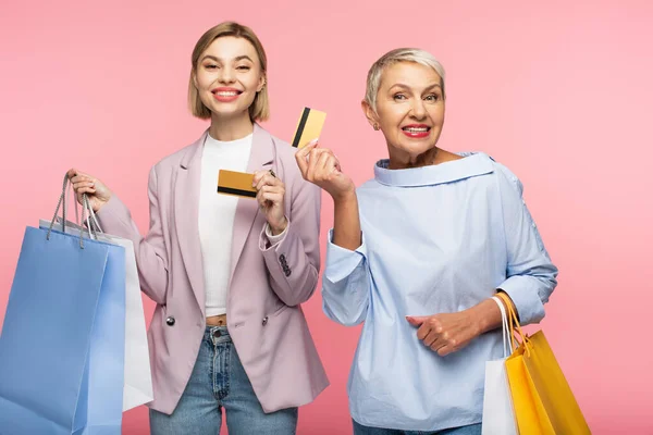 Glückliche junge Frau und lächelnde reife Mutter mit Einkaufstaschen und Kreditkarten isoliert auf rosa — Stockfoto