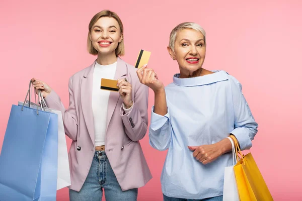 Feliz jovem e sorridente maduro mãe segurando sacos de compras e cartões de crédito isolados em rosa — Fotografia de Stock