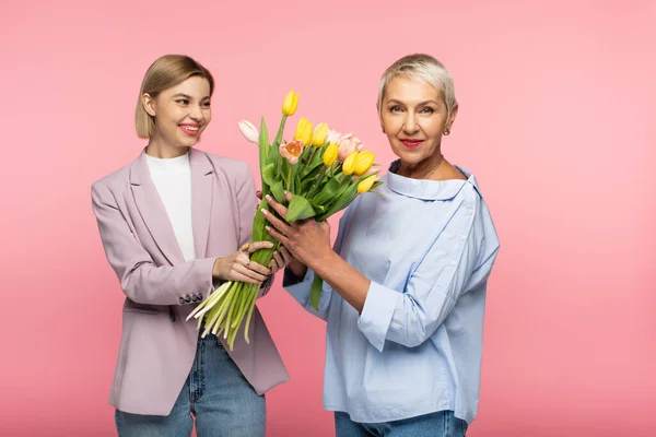 Glückliche junge Tochter hält Strauß Tulpen in der Nähe reife Mutter isoliert auf rosa — Stockfoto