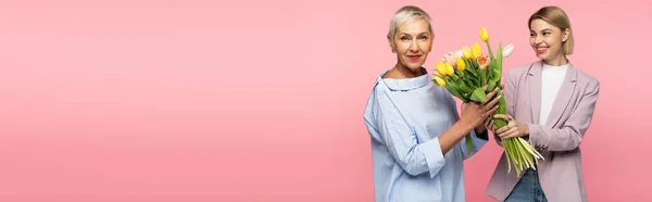 Happy young daughter holding bouquet of tulips near mature mother isolated on pink, banner — Stock Photo