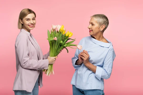 Glückliche junge Tochter gibt Blumenstrauß an fröhliche Mutter mittleren Alters isoliert auf rosa — Stockfoto