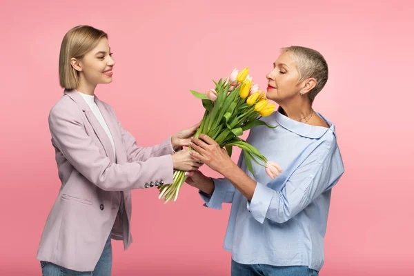 Erfreut kleine Tochter gibt Blumenstrauß an fröhliche Mutter mittleren Alters isoliert auf rosa — Stockfoto
