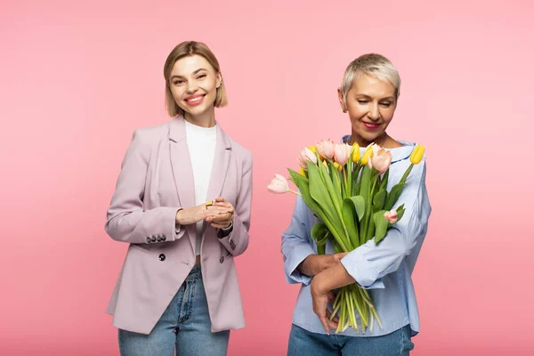 Feliz de mediana edad madre sosteniendo ramo de tulipanes cerca hija complacida aislado en rosa - foto de stock