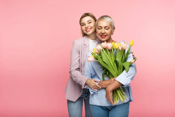 Zufriedene Tochter umarmt glückliche Mutter mittleren Alters hält Blumenstrauß isoliert auf rosa — Stockfoto