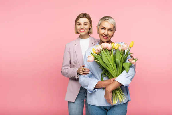 Fröhliche Tochter umarmt glückliche Mutter mittleren Alters hält Blumenstrauß isoliert auf rosa — Stockfoto