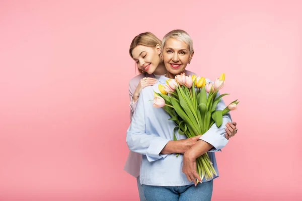 Alegre filha abraçando feliz meia idade mãe segurando tulipas isolado no rosa — Fotografia de Stock