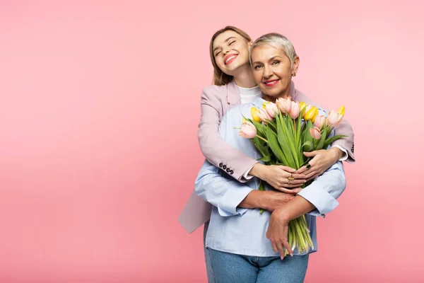 Positive daughter hugging happy middle aged mother holding tulips isolated on pink — Stock Photo