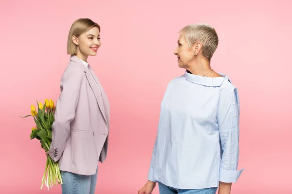 Hija joven positiva sosteniendo ramo de tulipanes detrás de la espalda cerca de la madre madura aislada en rosa - foto de stock