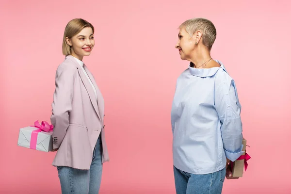Fröhliche junge Tochter und reife Mutter halten Geschenke hinter dem Rücken isoliert auf rosa — Stockfoto
