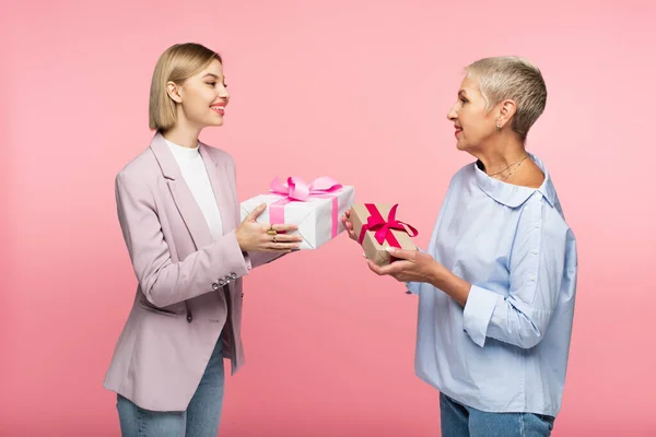 Alegre joven hija y maduro madre sosteniendo regalos aislado en rosa - foto de stock