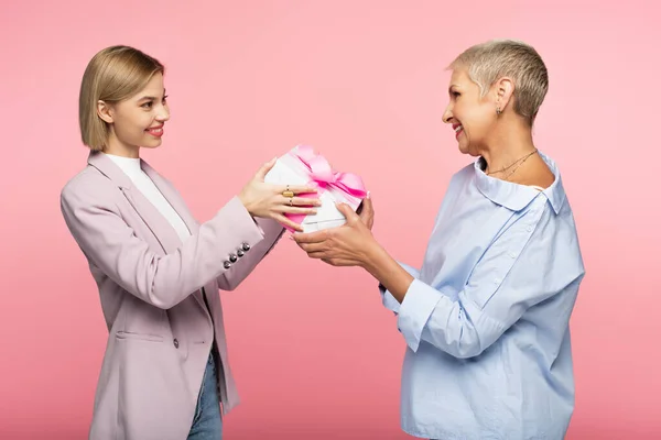 Glückliche Frau überreicht verpacktes Geschenk an fröhliche reife Mutter isoliert auf rosa — Stockfoto