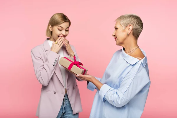 Зрелая мать дарит подарок изумленной маленькой дочери, изолированной на розовый — стоковое фото