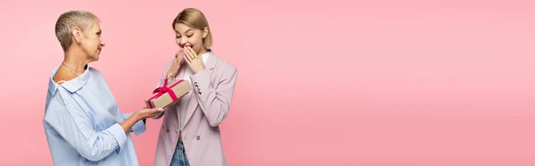 Mature mother giving present to amazed young daughter isolated on pink, banner — Stock Photo