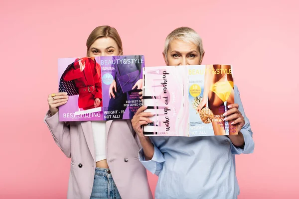 Зріла мати і молода дочка прикривають обличчя журналами ізольовані на рожевому — стокове фото