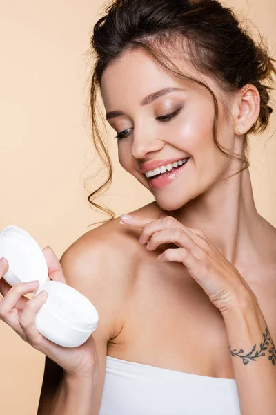 Mujer alegre aplicando crema cosmética en el hombro aislado en beige - foto de stock