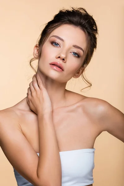 Pretty brunette woman looking at camera and touching neck isolated on beige — Stock Photo