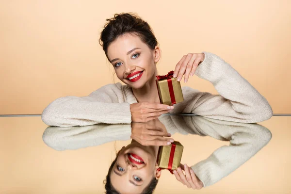 Усміхнена жінка з червоними губами тримає подарунок біля дзеркала ізольовано на бежевому — стокове фото
