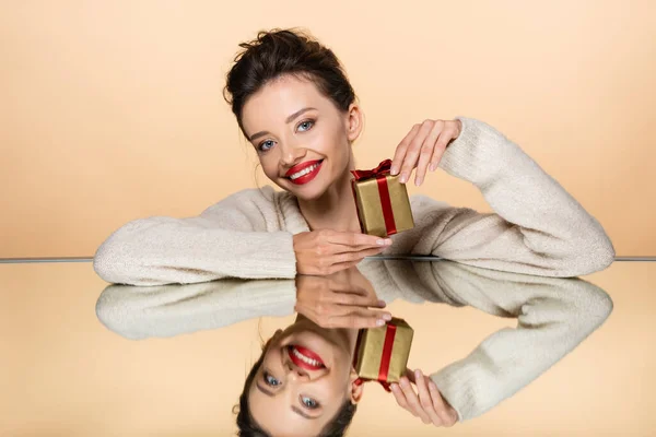 Mulher sorridente com lábios vermelhos segurando presente perto do espelho isolado no bege — Fotografia de Stock