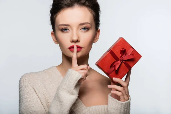 Donna in maglione in possesso di regalo rosso e mostrando gesto segreto isolato su bianco — Foto stock